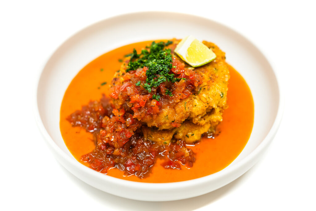 A plate of golden-brown crab cakes stacked atop a pool of creamy coconut curry sauce, topped with vibrant red bell pepper relish, garnished with fresh cilantro and a lime wedge.