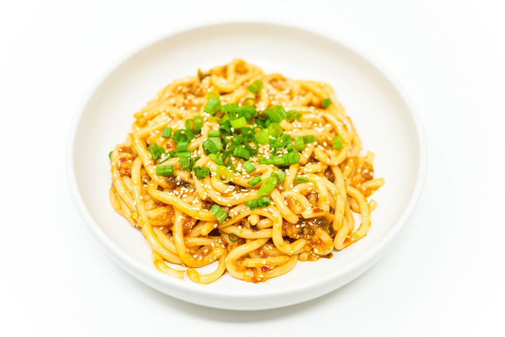 A bowl of Spicy Sesame Scallion Udon Noodles, featuring thick, chewy noodles coated in a rich, savory, and spicy sauce, garnished with fresh scallions and toasted sesame seeds.