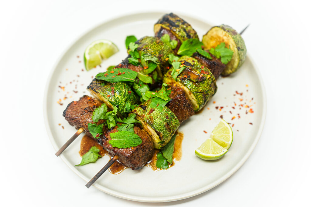 A platter of Tangy Grilled Beef and Zucchini Skewers, featuring marinated beef and zucchini chunks, drizzled with balsamic vinaigrette, and garnished with fresh mint, cilantro, parsley, and red pepper flakes, served with lime wedges on the side.