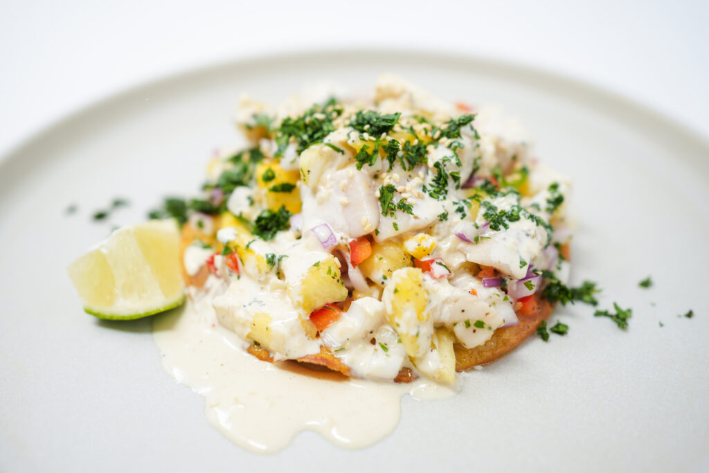 Swordfish and pineapple ceviche served on a crispy tostada, garnished with fresh cilantro and a lime wedge, drizzled with aioli for a vibrant and refreshing flavor combination.