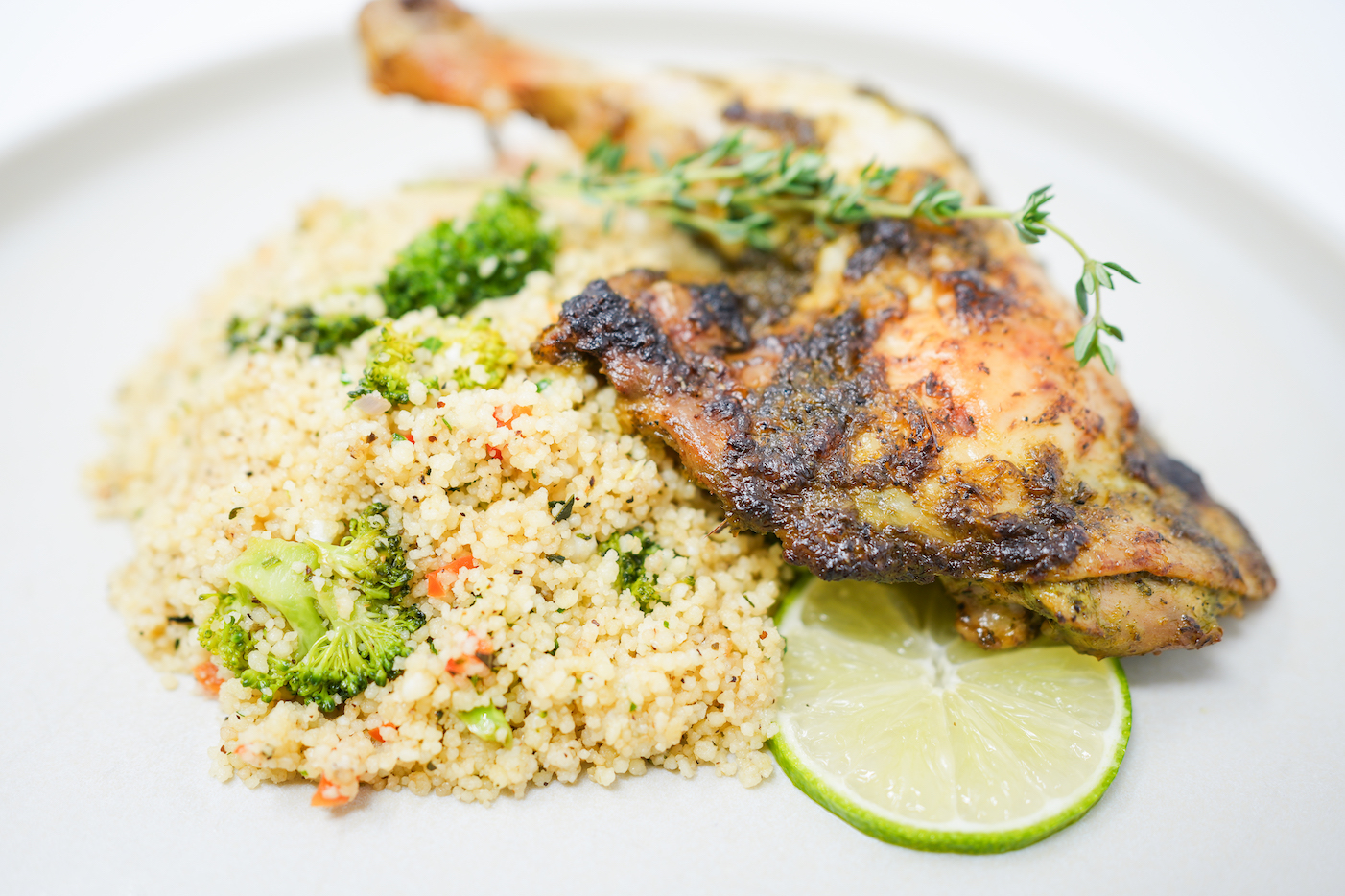A plate of Citrus Herb Roasted Chicken featuring golden-brown, crispy chicken pieces garnished with fresh herbs and lime slices, served alongside a vibrant broccoli couscous.
