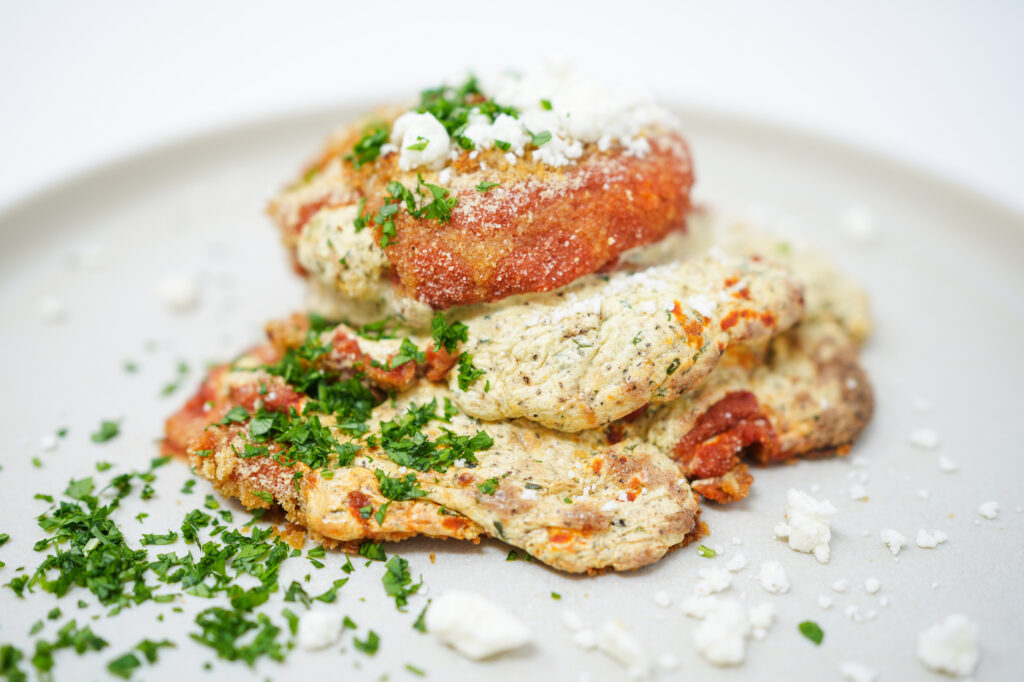 A plated dish of Fried Eggplant Ricotta Stacks, featuring layers of crispy fried eggplant slices, creamy ricotta cheese filling, and fresh tomato sauce, topped with crumbled goat cheese and freshly chopped parsley