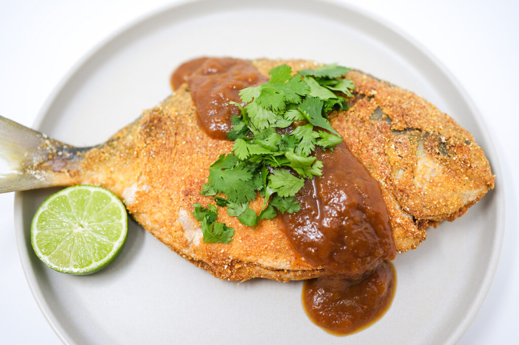 A beautifully plated dish of Coconut Curry Fried Pomfret, garnished with fresh cilantro and lime wedges, served alongside a tangy tamarind chutney. The fish is golden brown and crispy, with a vibrant and colorful presentation.