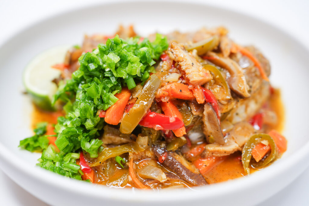 A vibrant plate of Kingfish Escovitch featuring crispy fried kingfish steaks topped with a tangy, spicy vegetable medley of carrots, onions, bell peppers, and Scotch bonnet peppers, garnished with chopped green onions and served with lime wedges.
