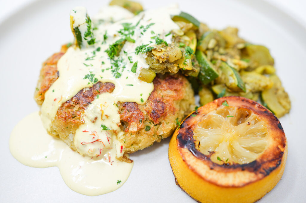 A plate of golden-brown pollock fish cakes drizzled with saffron aioli, served alongside colorful roasted yellow squash and zucchini seasoned with za'atar, garnished with fresh parsley and caramelized lemon slices.