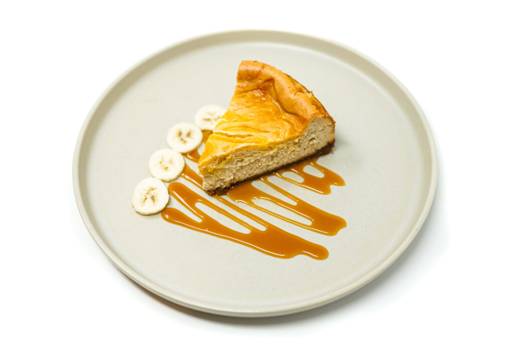 Slice of banana mango swirl cheesecake with a graham cracker crust, served on a plate with caramel sauce and banana slices.