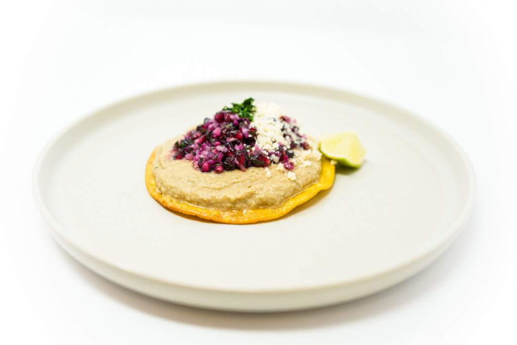 A crispy tostada topped with creamy baba ganoush, vibrant pomegranate salsa, crumbled Cotija cheese, and fresh cilantro, served with lime wedges.