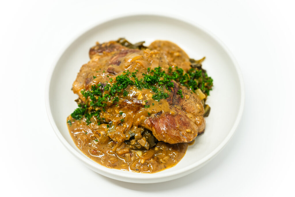 A plate of Smothered Pork Butt Steaks served on top of beer-braised collard greens, topped with rich gravy and garnished with fresh parsley.