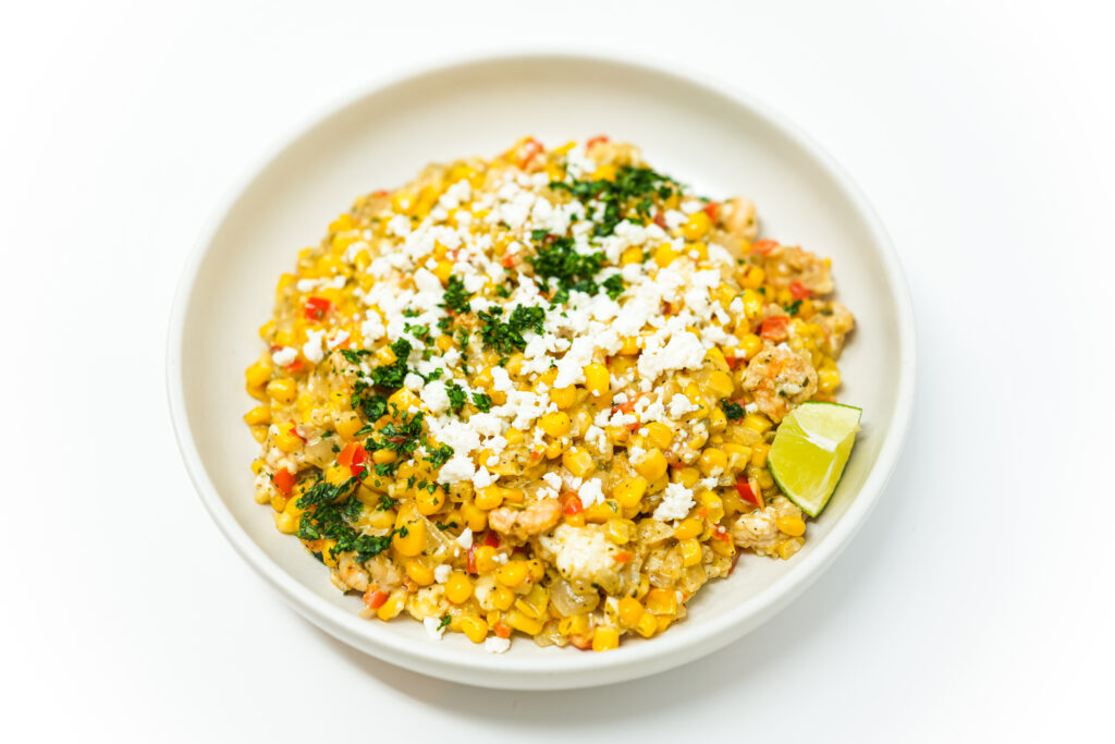 A vibrant Shrimp and Corn Esquites featuring charred corn, tender shrimp, and a creamy sauce, garnished with crumbled cotija cheese and fresh cilantro, with lime wedges on the side.