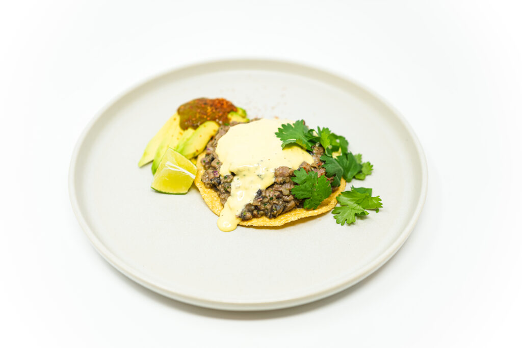 A plate of Mexican Fusion Portobello Mushroom Beef Tartare, topped with fresh cilantro and avocado slices, served alongside crispy corn tortillas and drizzled with garlic aioli for a vibrant, modern presentation.