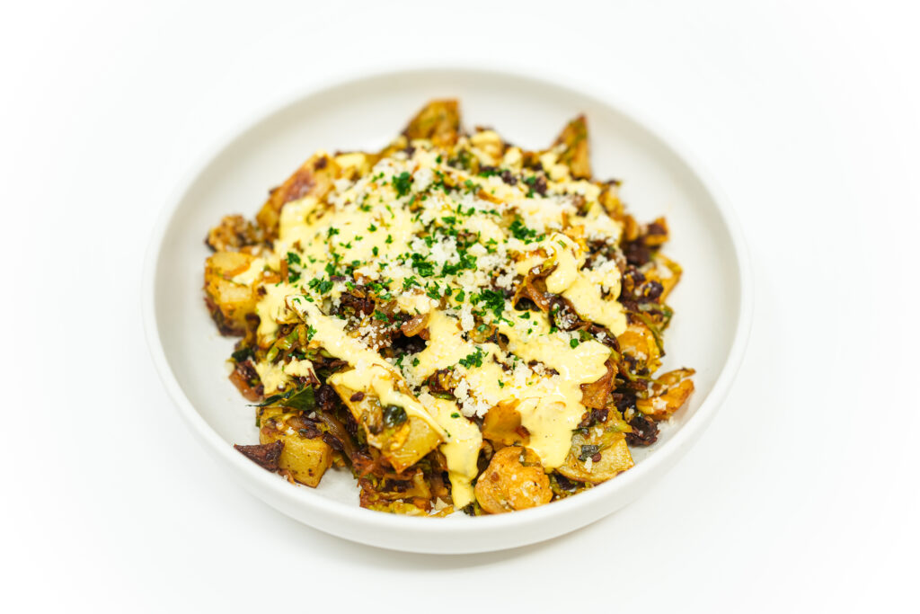 Brussels Sprouts and Sun-Dried Tomato Hash served on a plate, featuring crispy potatoes, caramelized Brussels sprouts, and tangy sun-dried tomatoes, drizzled with creamy tahini aioli and garnished with Parmesan cheese and fresh parsley