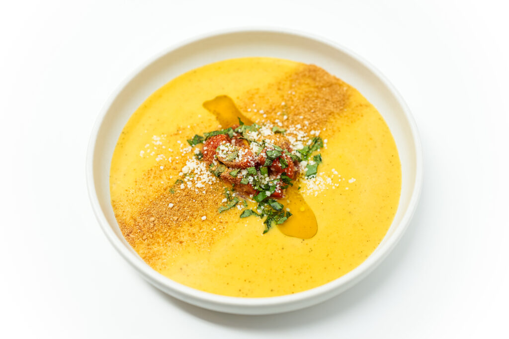 A bowl of silky yellow tomato gazpacho garnished with chopped basil, toasted breadcrumbs, and grated Parmesan cheese, showcasing the soup's vibrant golden color and smooth, creamy texture.