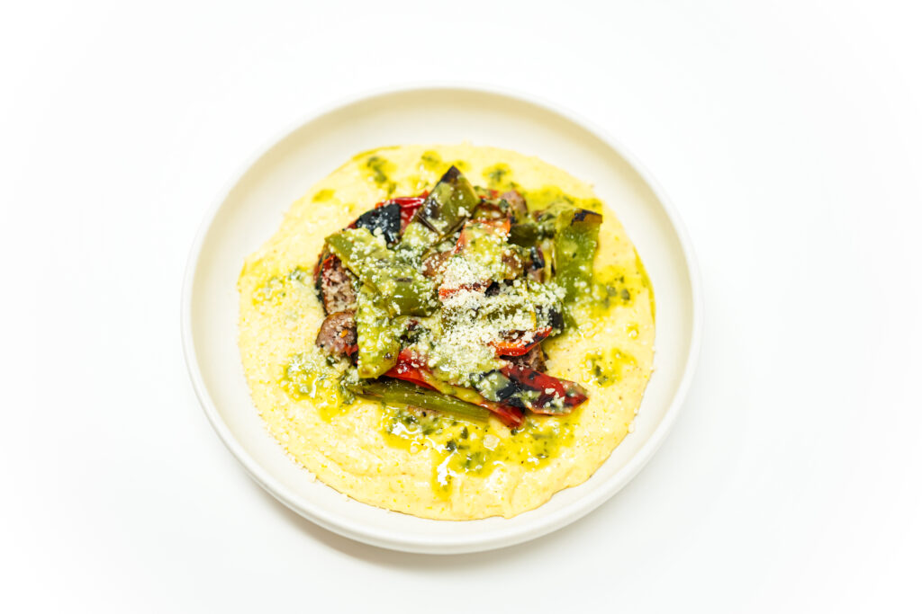 A plate of Herb Grilled Sausage and Peppers served over creamy Parmesan polenta, drizzled with a vibrant herb vinaigrette, and garnished with fresh herbs and grated Parmesan cheese.