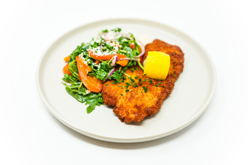 A plated dish of Crispy Chicken Milanese with golden-brown breaded chicken cutlets topped with a vibrant arugula salad, featuring roasted red peppers, thinly sliced red onion, and a sprinkle of Parmesan cheese, garnished with a lemon wedge and fresh parsley.
