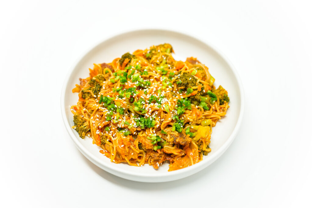 A bowl of Broccoli and Bacon Yaki Udon featuring thick udon noodles stir-fried with crispy bacon, tender broccoli, and vibrant vegetables, garnished with chopped green onions and sesame seeds.