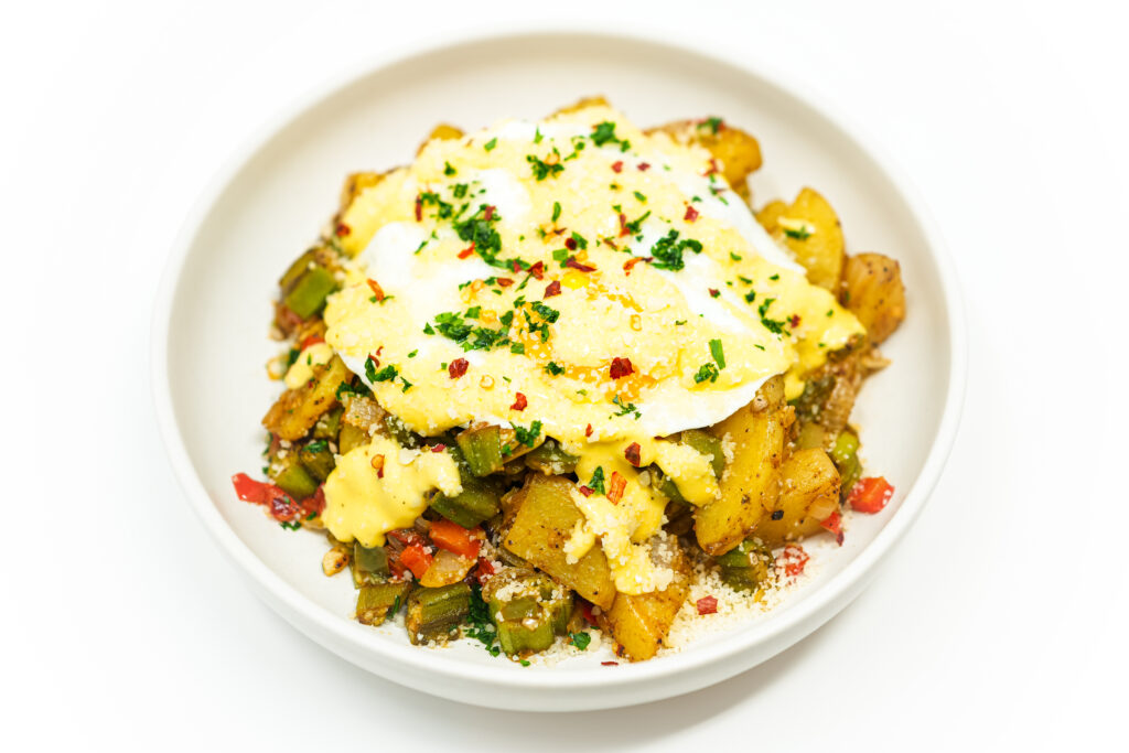 Southern okra hash with crispy potatoes, sautéed vegetables, a fried egg, and creamy tahini aioli, garnished with parsley and chili flakes.