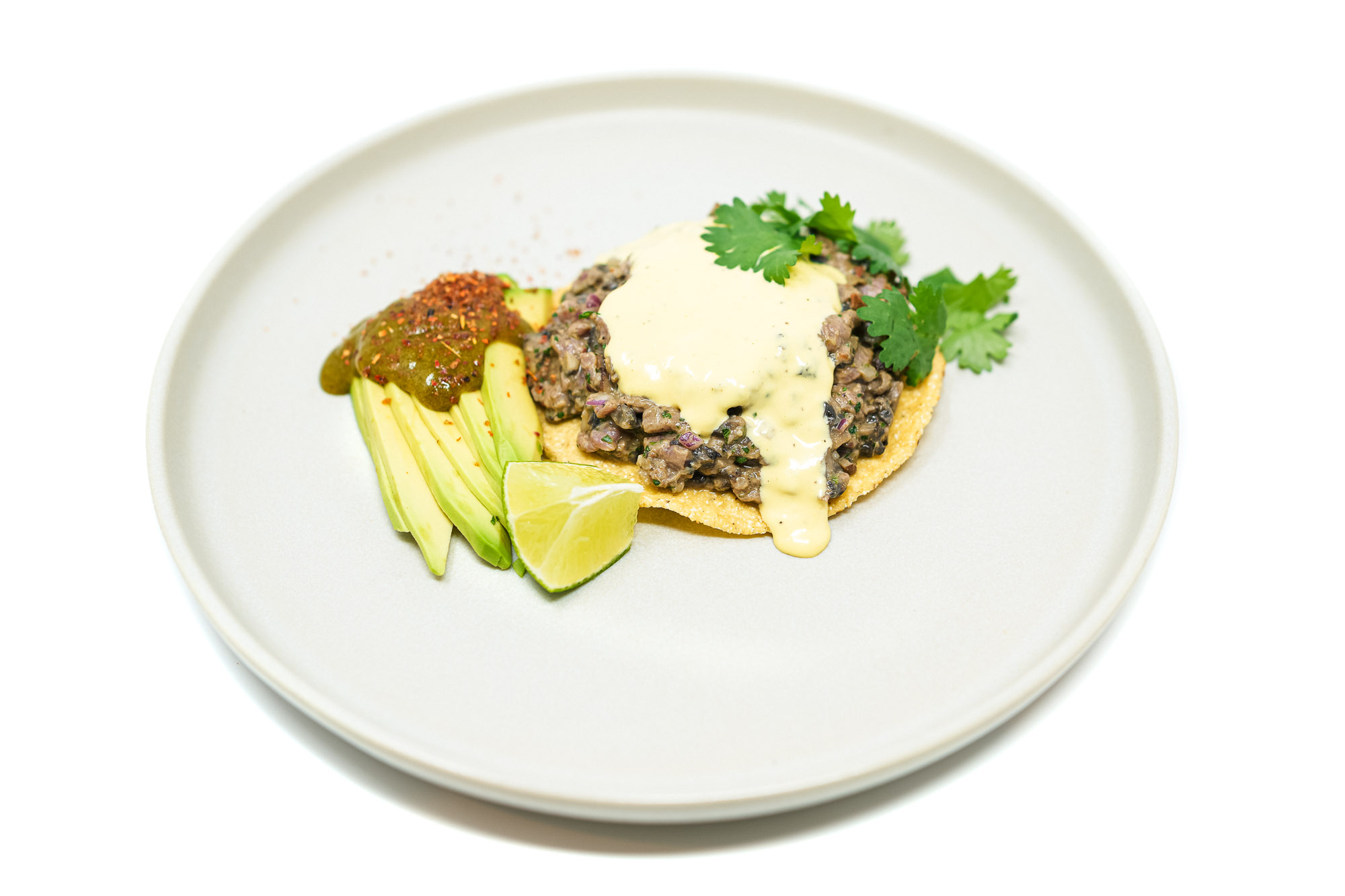 Close-up angled shot of hand-cut beef tenderloin and portobello mushroom tartare piled on a crisp round tostada, draped with creamy garlic aioli and served with sliced avocado and a fresh lime wedge on a light gray plate.