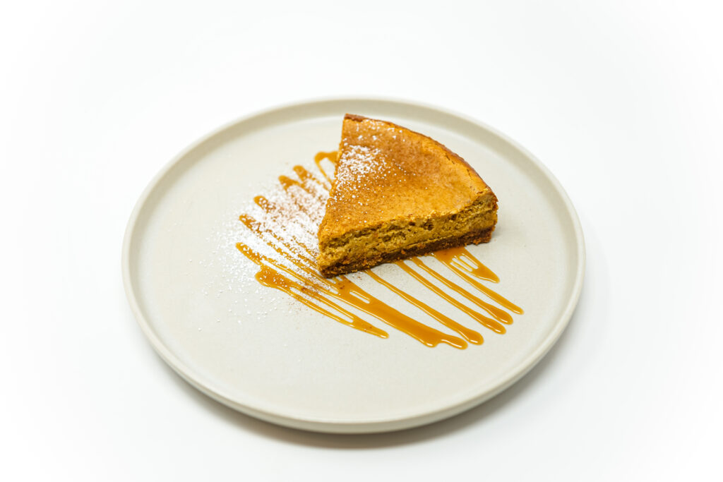 A slice of pumpkin cheesecake with a brown butter graham cracker crust, garnished with a drizzle of caramel and a sprinkle of powdered sugar, sitting on a plate for a festive fall dessert.