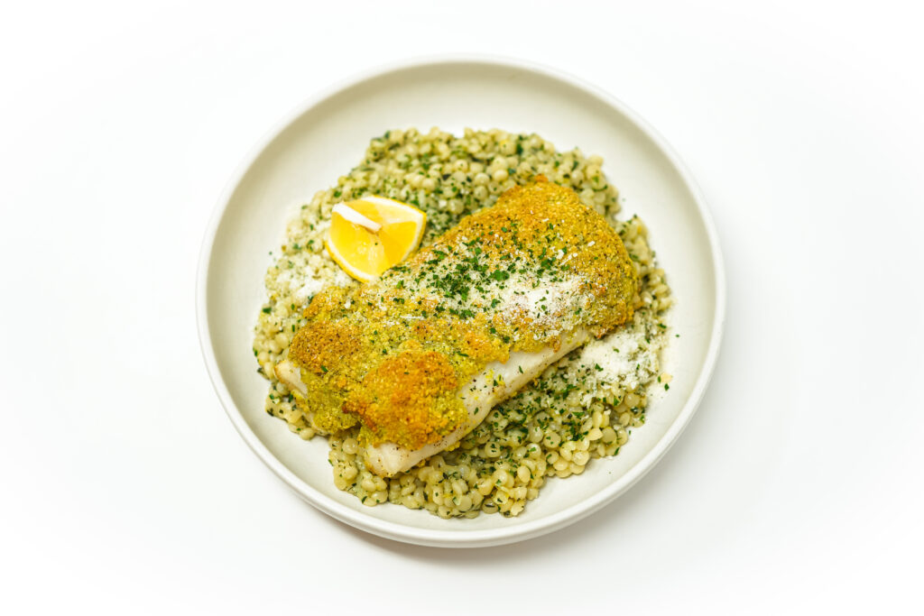 Pistachio-crusted cod fillet served on a bed of lemon herb pearl couscous, garnished with fresh herbs and a lemon wedge.