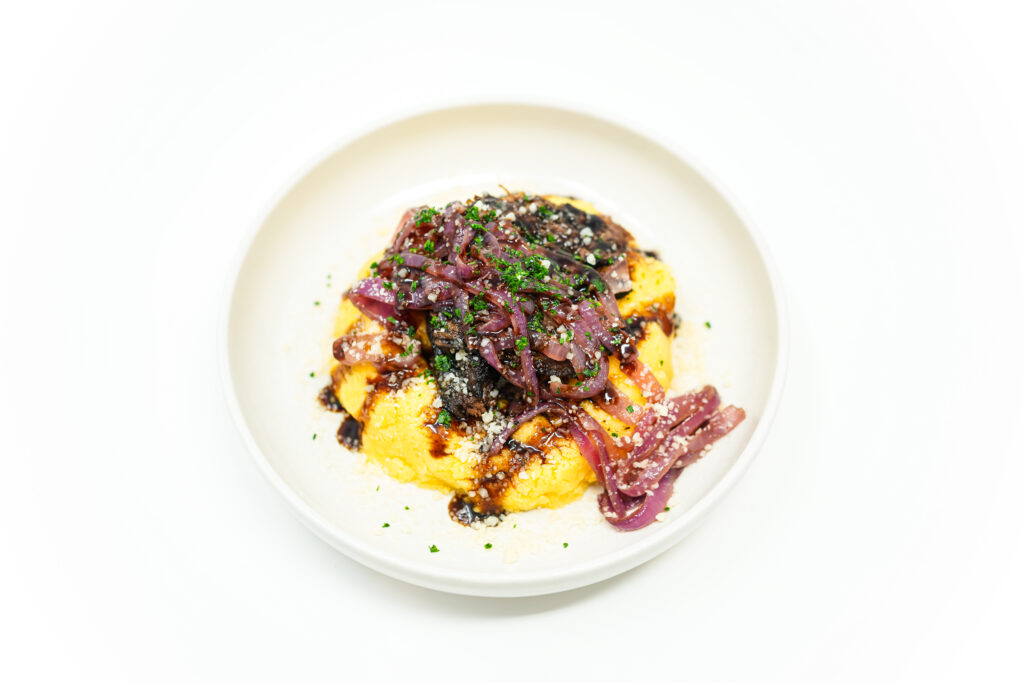 A plate of tender shredded brisket served over creamy Parmesan polenta, topped with sweet and tangy caramelized balsamic red onions. The dish is garnished with freshly chopped parsley and a drizzle of balsamic glaze, with a sprinkle of Parmesan cheese for added flavor.