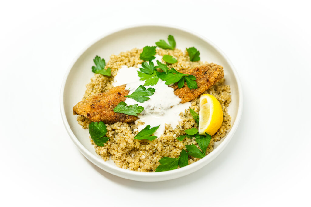 A plated dish of crispy pan-fried Spanish mackerel topped with creamy tahini aioli, served alongside fluffy garlic butter couscous, garnished with fresh herbs and a lemon wedge.