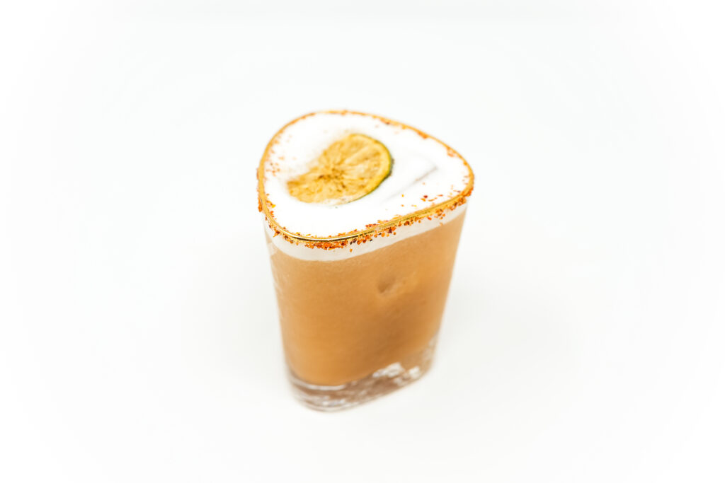 A Tamarind Pineapple Tequila Sour served in a glass, topped with a frothy layer and garnished with a smoky charred lime wheel, set against a white background.