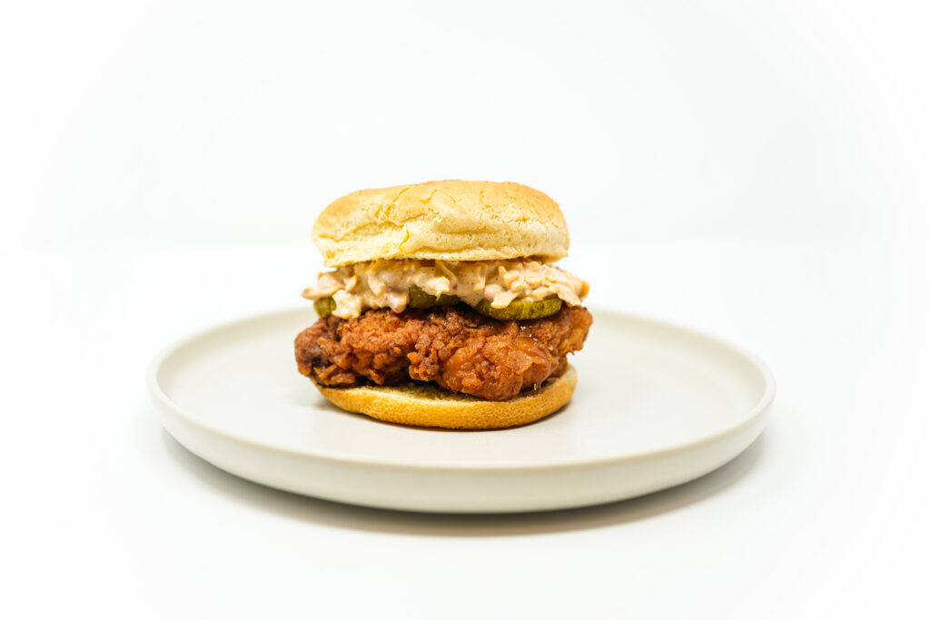 Hot Honey Chicken Pimento Cheese Sandwich with crispy fried chicken, creamy pimento cheese, pickle slices, and a drizzle of hot honey on a brioche bun.