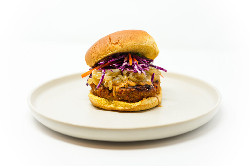 A Cajun Shrimp Burger featuring a golden, spiced shrimp patty topped with caramelized onions, vibrant purple cabbage slaw, and a drizzle of smoky remoulade, served on a toasted brioche bun.