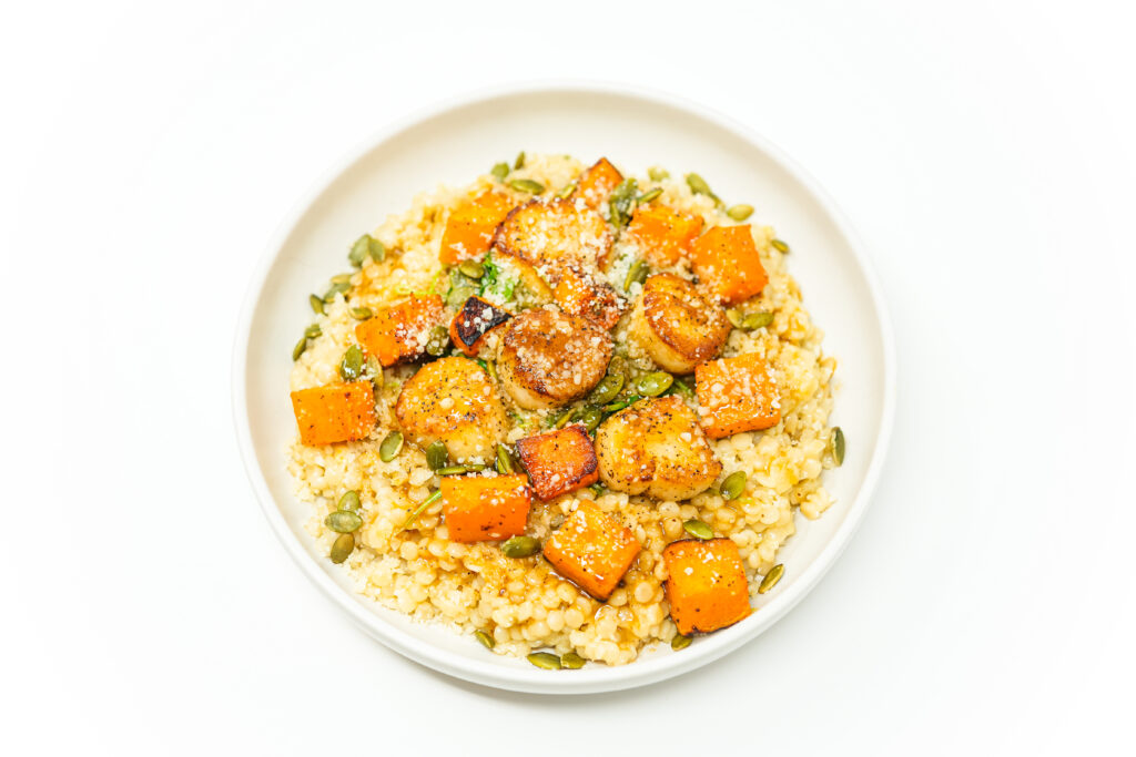 A beautifully plated dish featuring golden seared scallops atop layers of pearl couscous, roasted butternut squash, and fresh arugula, drizzled with champagne brown butter sauce and garnished with toasted pumpkin seeds and Parmesan cheese.