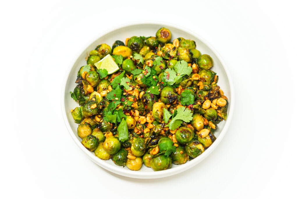 A vibrant plate of roasted Brussels sprouts drizzled with Thai-inspired mustard sauce, garnished with roasted peanuts, fresh cilantro, and lime wedges for a bold and colorful presentation