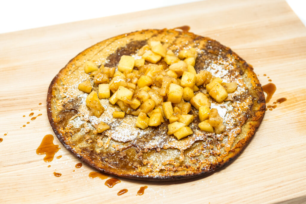 A golden, puffy Caramel Apple Dutch Baby Pancake served in a cast-iron skillet, topped with tender caramelized apple slices and drizzled with rich caramel sauce, garnished with powdered sugar