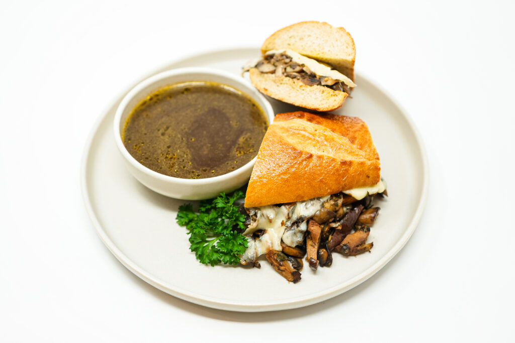 A Shiitake and Cremini Mushroom French Dip sandwich on a toasted baguette with melted Swiss cheese, served alongside a bowl of rich soy-based broth for dipping.