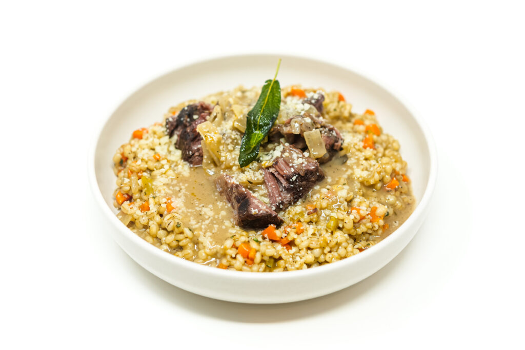 High-angle shot of a white bowl filled with pearl barley and topped with tender chunks of braised short ribs, covered in a light brown sage gravy and garnished with a single fresh sage leaf.