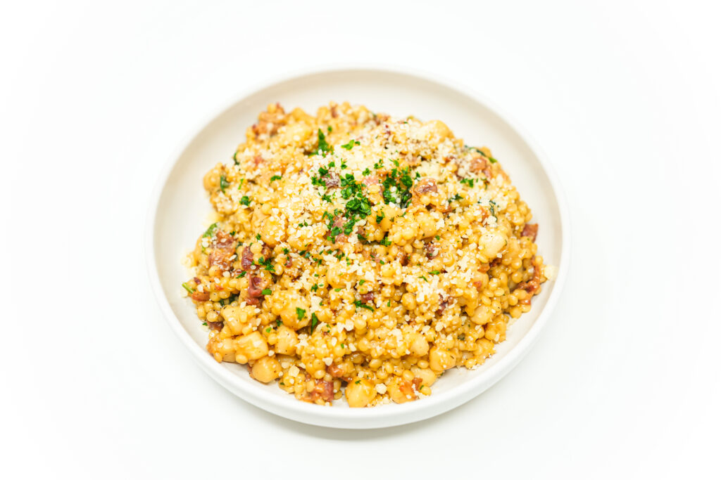 Creamy pearl couscous with tender bay scallops, crispy bacon, fresh parsley, and Parmesan cheese, beautifully plated in a white bowl on a clean, minimalist background.