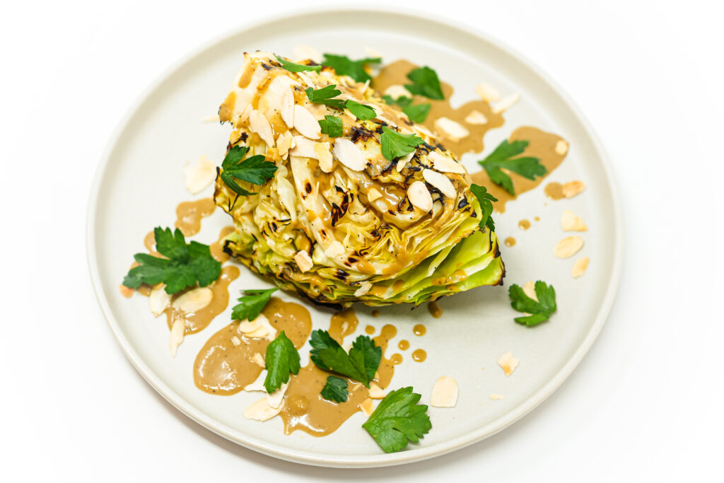 Grilled cabbage wedge drizzled with a Thai-inspired mustard sauce, garnished with toasted almonds and fresh cilantro, served on a white plate.