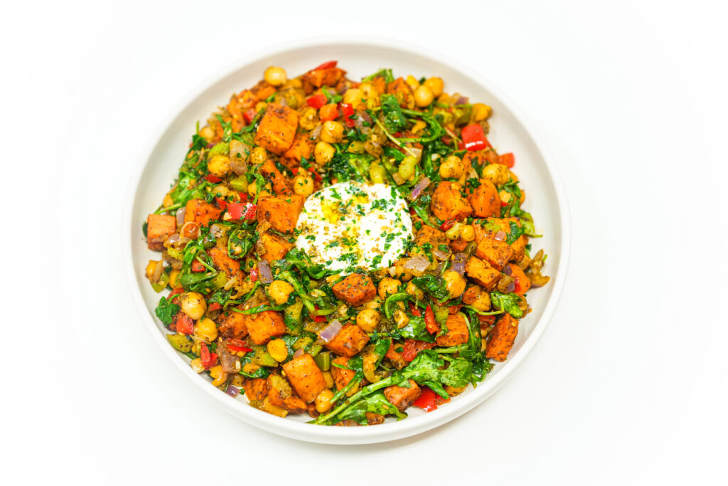 A vibrant bowl of sweet potato and chickpea hash with roasted sweet potatoes, chickpeas, red bell peppers, arugula, and a dollop of yogurt, garnished with fresh parsley.