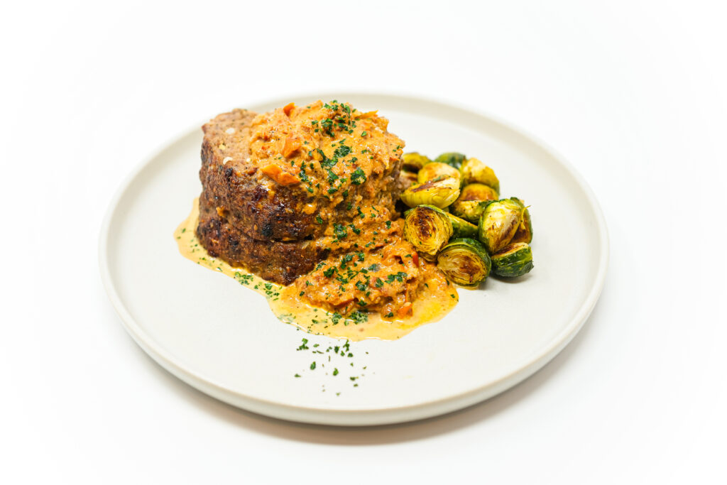 A plate featuring Italian Sausage Meatloaf topped with a creamy sun-dried tomato sauce, served alongside roasted Brussels sprouts, garnished with fresh parsley.