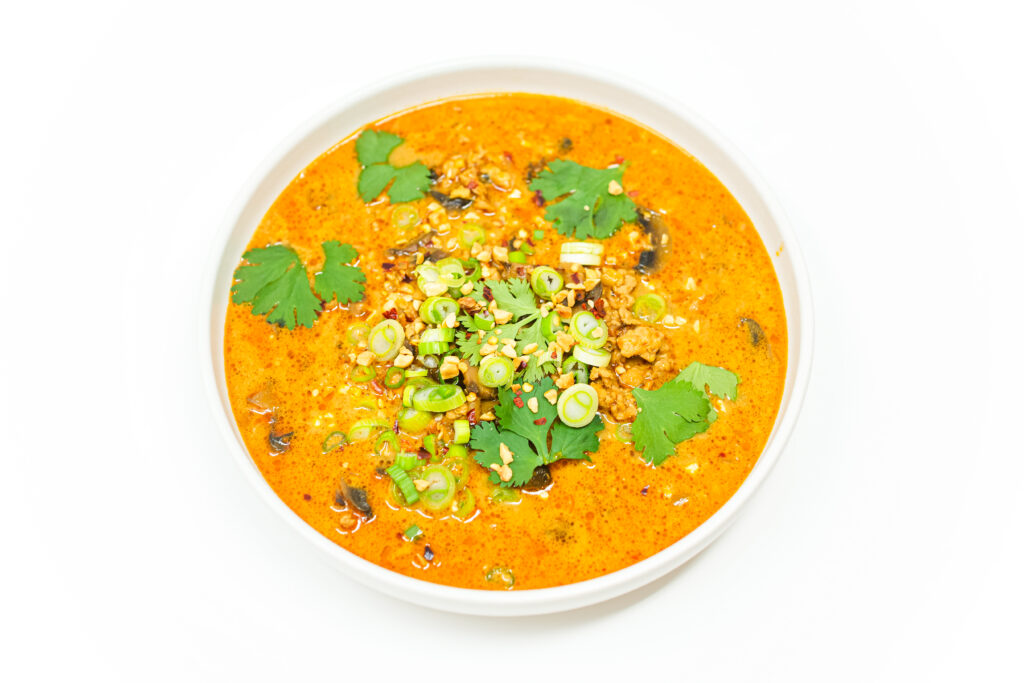 A bowl of Thai-Inspired Ground Chicken and Mushroom Soup, featuring a creamy peanut butter-infused broth, garnished with fresh cilantro, green onions, crushed peanuts, and red pepper flakes, served with lime wedges on the side.