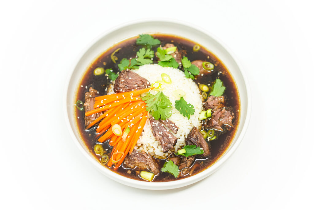 A bowl of Asian-Inspired Oxtail Soup with Rice, featuring tender braised oxtail in a rich, aromatic broth with jasmine rice, pickled carrots, fresh cilantro, green onions, and sesame seeds, served in a white bowl.