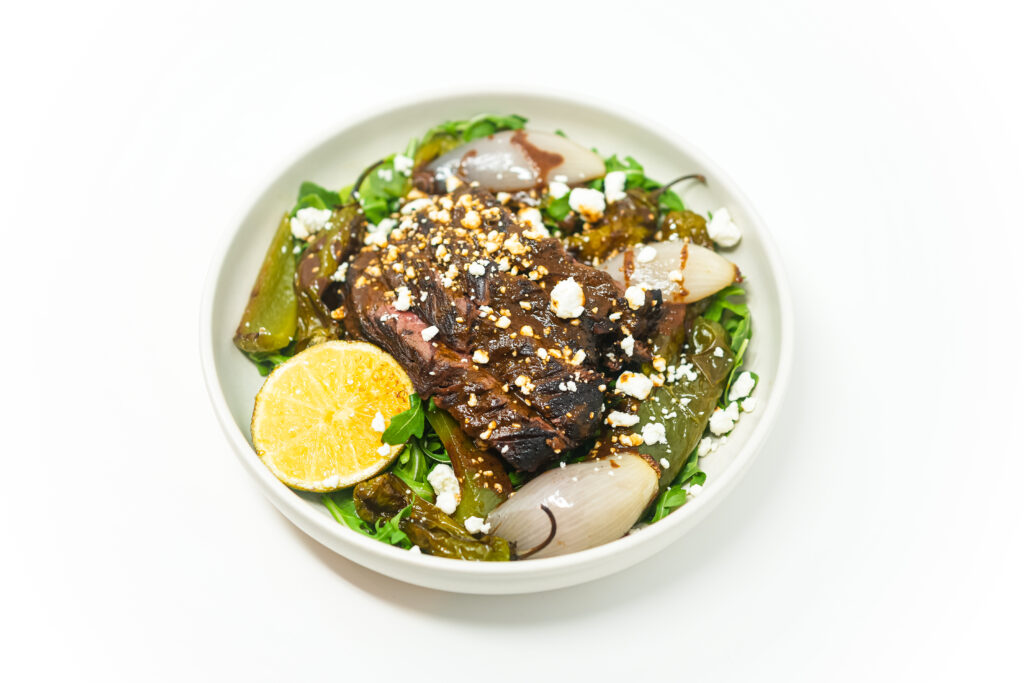 A plate of Soy-Lime Grilled Hanger Steak and Peppers, featuring sliced hanger steak with a caramelized crust, charred shishito and green bell peppers, grilled shallots, and crumbled goat cheese, served over arugula with grilled limes on the side. A drizzle of reduced soy-lime glaze adds a rich, umami-packed finish.