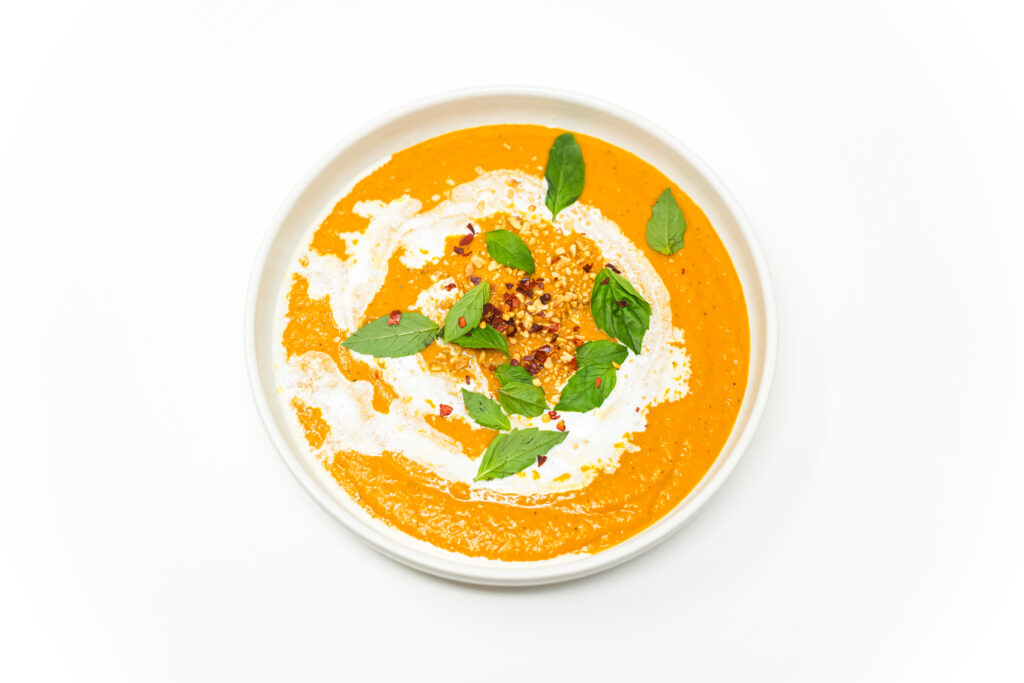A bowl of Thai-Inspired Butternut Squash and Rutabaga Soup, garnished with a swirl of coconut cream, fresh Thai basil, sesame seeds, and red pepper flakes. The soup’s rich golden-orange color contrasts with the vibrant toppings, creating a warm and inviting presentation.