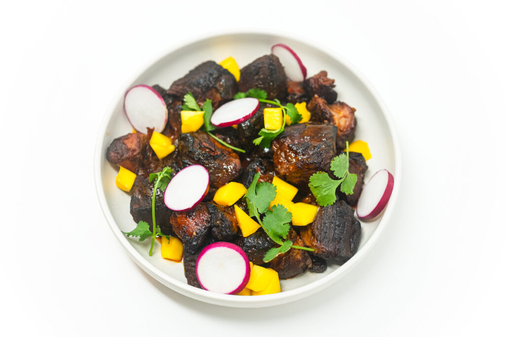 A plate of crispy mango and makrut lime pork carnitas, garnished with fresh cilantro, thinly sliced radishes, and diced mango, served in a white bowl.