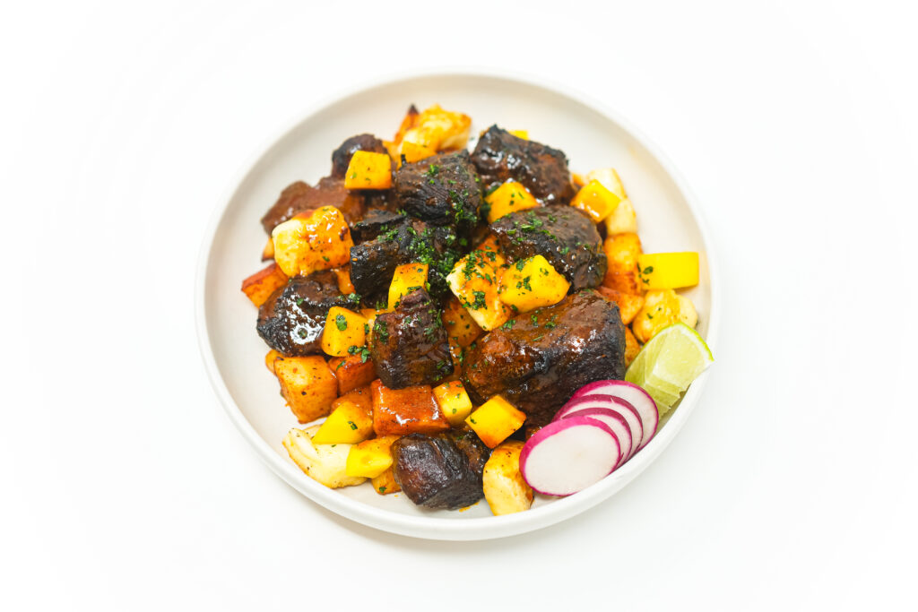 A plate of Mango and Makrut Lime Carnitas served with crispy paprika-roasted potatoes, golden-brown fried paneer cheese, and fresh garnishes of diced mango, sliced radishes, and chopped cilantro.