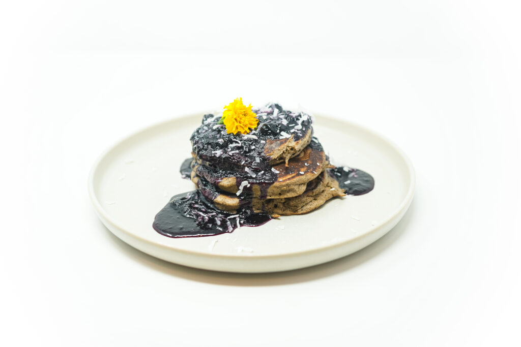 A stack of fluffy buckwheat pancakes topped with deep purple blueberry syrup, dusted with coconut flakes, and garnished with an edible yellow flower, served on a minimalist white plate.