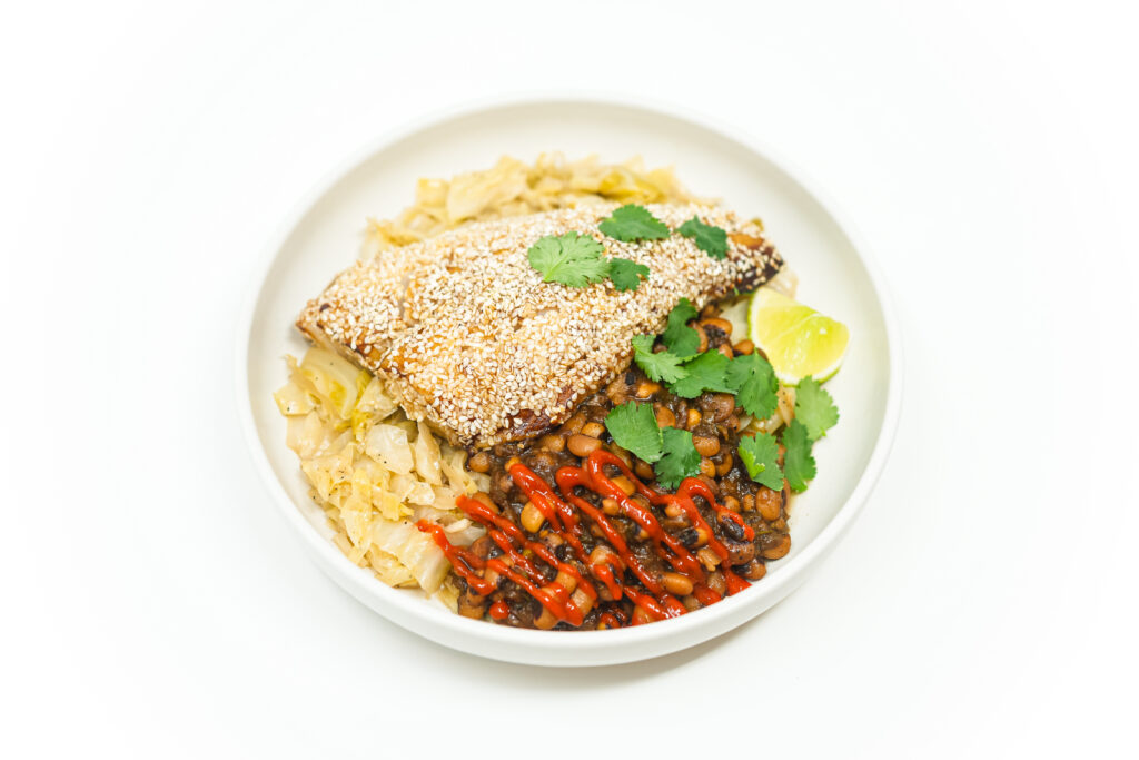 A beautifully plated dish of Sesame-Crusted Black Cod with a golden, crispy sesame seed crust, served over soy-braised cabbage and spicy black-eyed peas, garnished with fresh cilantro and lime wedges.
