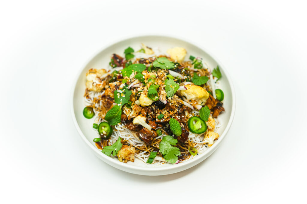 A bowl of hoisin-glazed chicken and roasted cauliflower served over vermicelli noodles, garnished with fresh cilantro, mint, jalapeños, sesame seeds, and crushed peanuts. The dish is drizzled with a savory hoisin sauce and presented in a clean, minimalist setting.