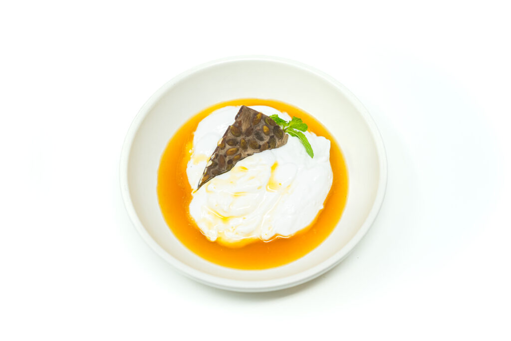 A creamy Mango Coconut Mousse served in a glass dish, topped with silky mango sauce, a shard of pumpkin seed chocolate bark, and fresh mint, set against a bright, neutral background.