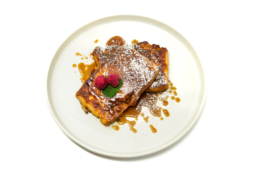 A close-up image of Coconut Curry Naan French Toast, featuring golden-brown pieces of spiced naan topped with a rich coconut caramel sauce, garnished with powdered sugar and a sprinkle of cinnamon, served with fresh berries.