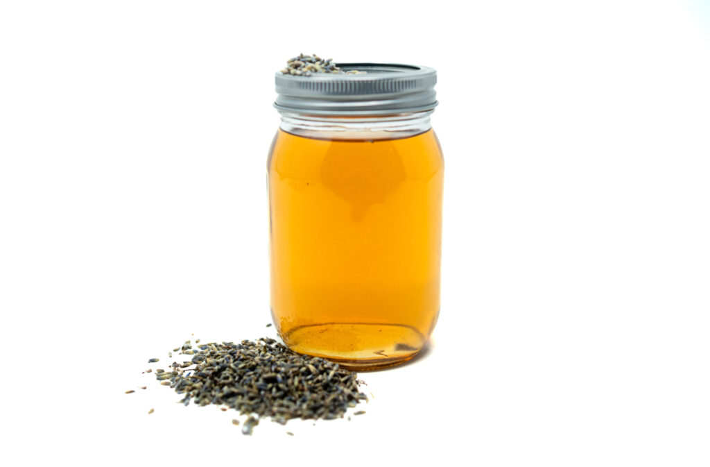 A glass jar filled with golden lavender-infused vodka, placed on a white background with dried lavender buds scattered beside it.