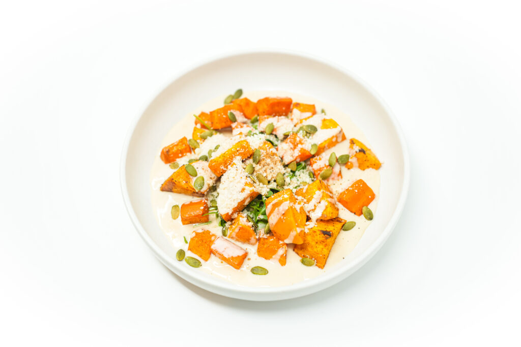 Charred sweet potatoes and carrots plated on a creamy chai-spiced sauce, garnished with Parmesan, toasted pumpkin seeds, and fresh arugula.